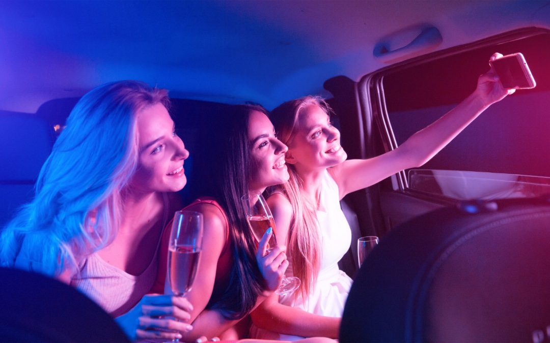 Three women in a car at night, smiling and taking a selfie while holding glasses of champagne.